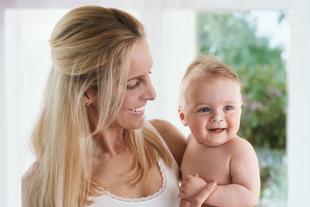 Eine Frau mit blondem Haar hält ein lächelndes Baby vor dem Hintergrund eines hellen Raums mit einem Fenster.