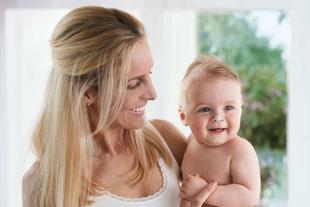 Eine Frau mit blondem Haar hält ein lächelndes Baby vor dem Hintergrund eines hellen Raums mit einem Fenster.