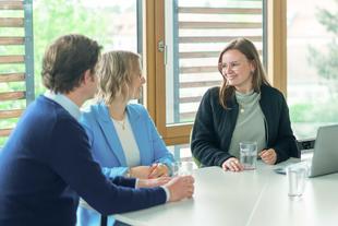 Drei Personen sitzen an einem Tisch vor einem Fenster und unterhalten sich, zwei Frauen und ein Mann, alle mit Brille und legerer Kleidung