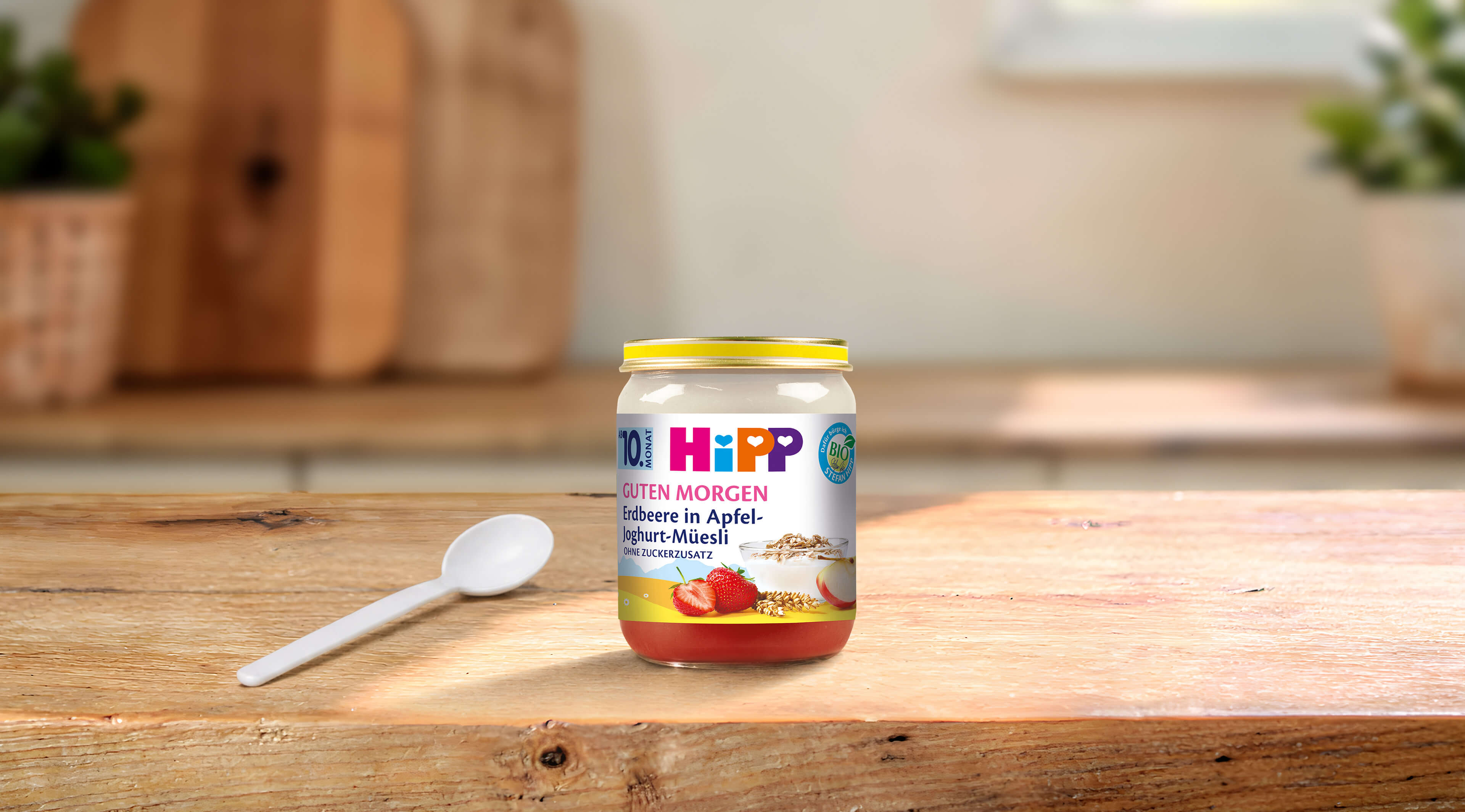 HiPP Guten Morgen Erdbeere in Apfel-Joghurt-Müesli Bio-Gläschen mit Löffel auf einem Holztisch in heller Küche