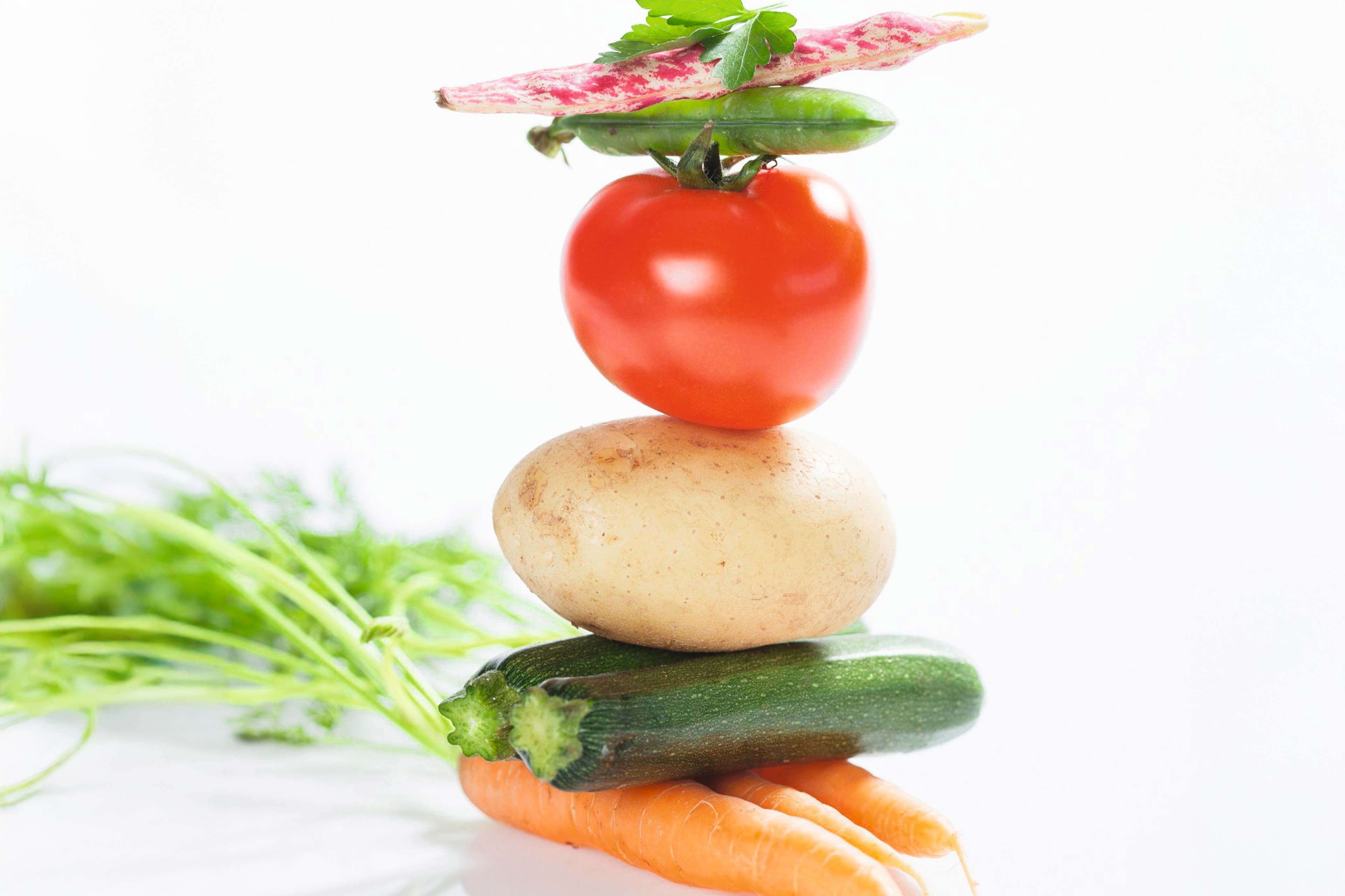 Ein Stapel frisches Gemüse, darunter eine Tomate, eine Kartoffel, eine Zucchini und eine Karotte, mit einem Hauch von grünen Kräutern im Hintergrund.