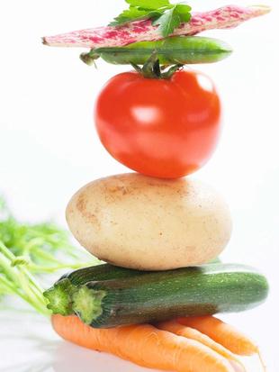Ein Stapel frisches Gemüse, darunter eine Tomate, eine Kartoffel, eine Zucchini und eine Karotte, mit einem Hauch von grünen Kräutern im Hintergrund.