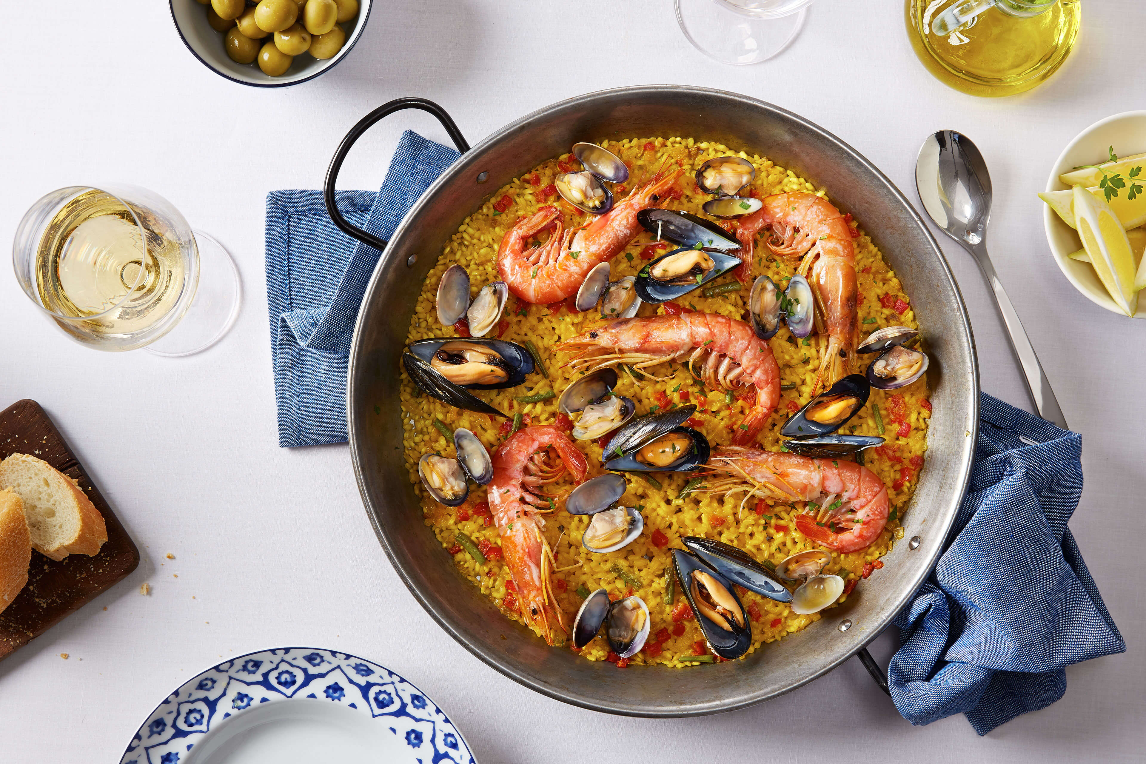 Ein Paella-Gericht mit Krabben, Muscheln und anderen Zutaten, begleitet von einem Glas Weißwein, einer Scheibe Brot und einigen Gewürzen.