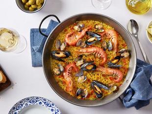 Ein Paella-Gericht mit Krabben, Muscheln und anderen Zutaten, begleitet von einem Glas Weißwein, einer Scheibe Brot und einigen Gewürzen.