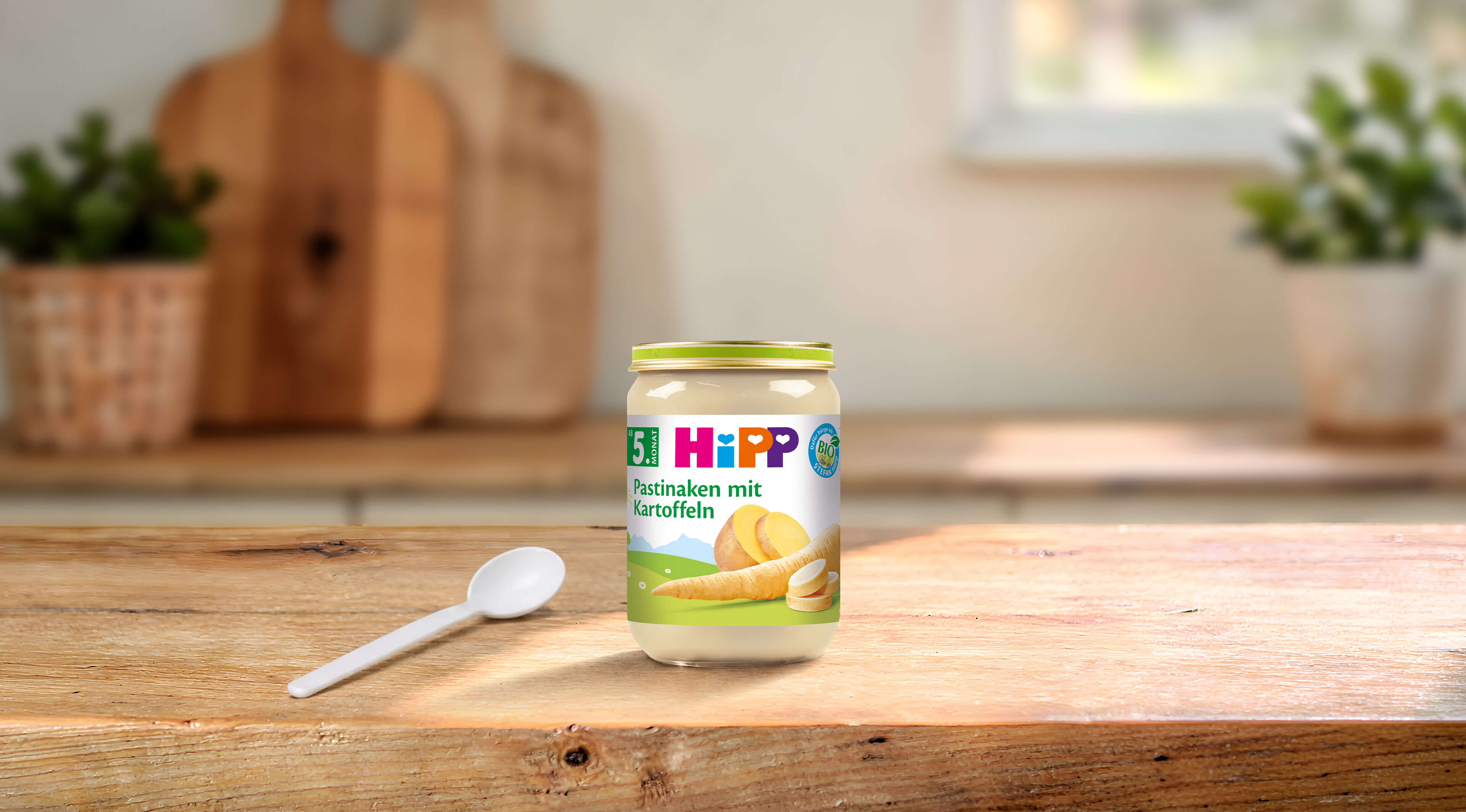 HiPP Bio-Gläschen „Pastinaken mit Kartoffeln“ ab dem 5. Monat mit Löffel auf Holztisch in heller Küche