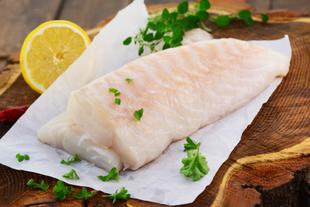 Ein Stück roher Fisch auf weißem Papier, garniert mit grünen Kräutern und einer Zitronenscheibe, auf einer Holzunterlage platziert.