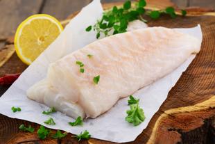 Ein Stück roher Fisch auf weißem Papier, garniert mit grünen Kräutern und einer Zitronenscheibe, auf einer Holzunterlage platziert.