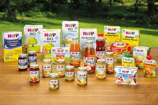 Verschiedene HiPP Bio-Babynahrung, Säfte und Kindermilch stehen bunt arrangiert auf einem Holztisch vor grüner Naturkulisse.