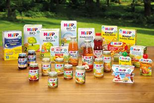 Verschiedene HiPP Bio-Babynahrung, Säfte und Kindermilch stehen bunt arrangiert auf einem Holztisch vor grüner Naturkulisse.