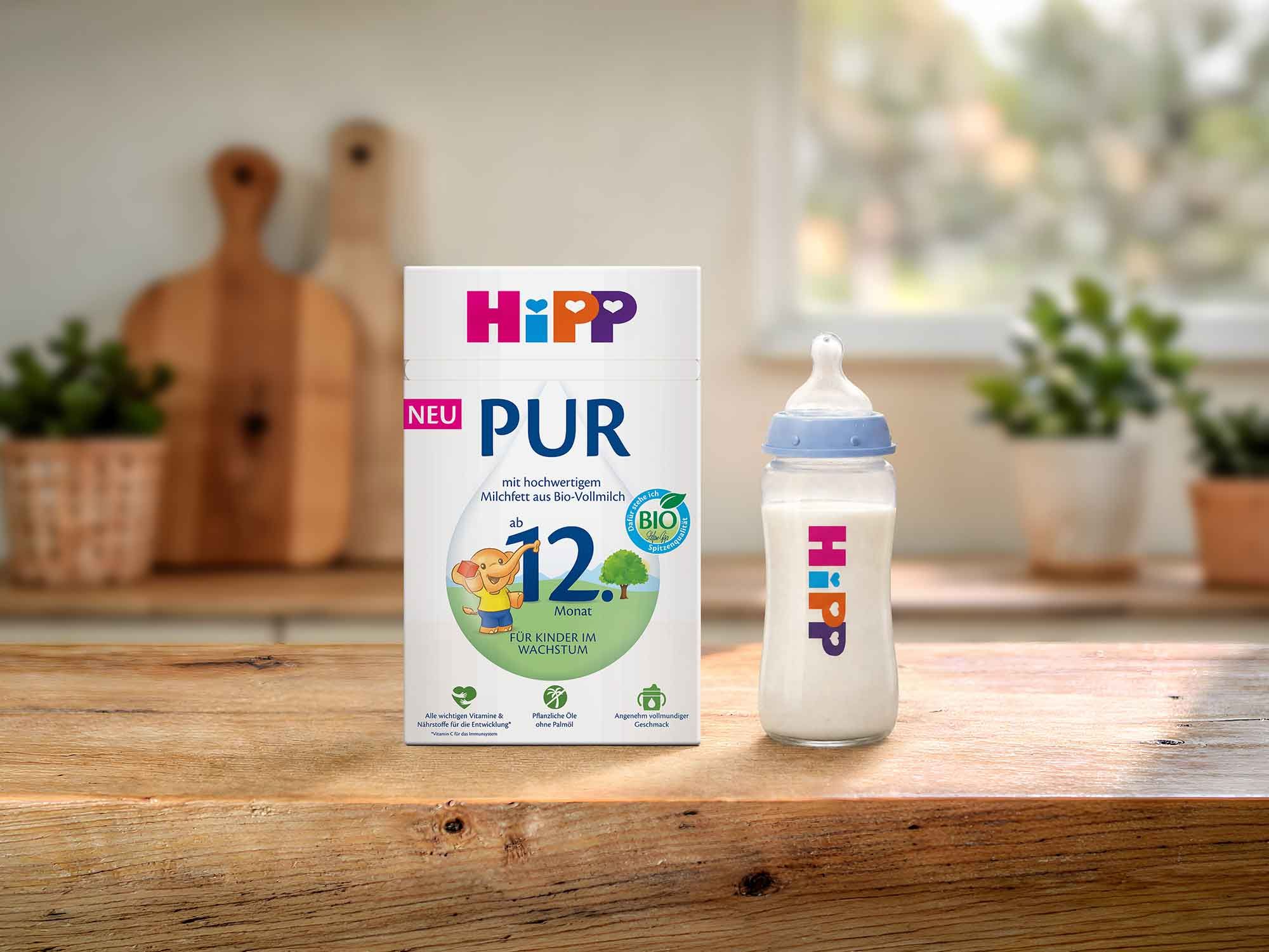 HiPP Kindermilch PUR Packung und gefülltes Fläschchen auf Holztisch in heller Küche, geeignet ab 12 Monaten