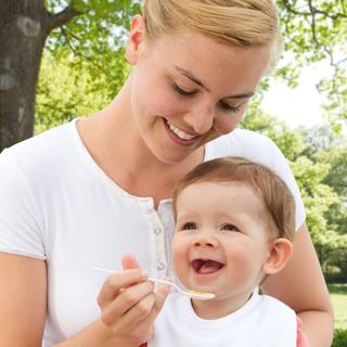 Eine Frau füttert ein Baby mit einem Löffel vor einer grünen Kulisse.