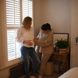 Two people stand by a bright window with white shutters in a cozy bedroom. One person in a white top and jeans leans casually on the windowsill while the other, dressed in a beige outfit, gently touches their belly in a warm, affectionate gesture.