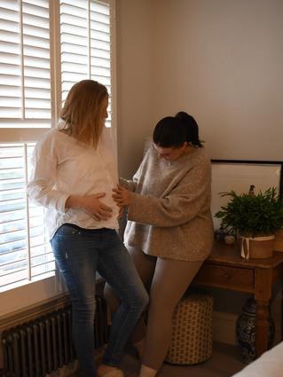 Two people stand by a bright window with white shutters in a cozy bedroom. One person in a white top and jeans leans casually on the windowsill while the other, dressed in a beige outfit, gently touches their belly in a warm, affectionate gesture.