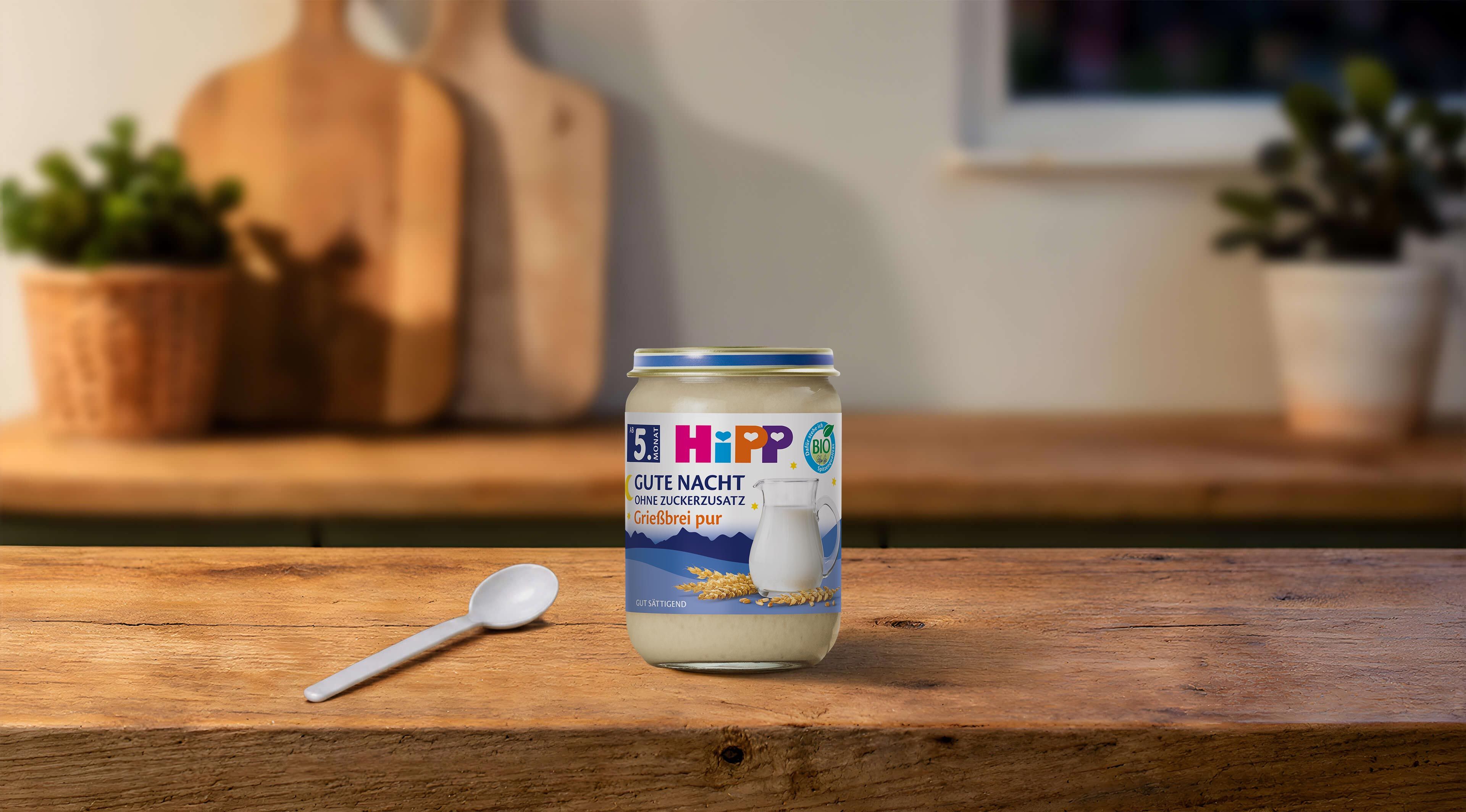 HiPP Gute Nacht Grießbrei pur im Glas auf einem Holztisch, mit Babylöffel davor in einer warm beleuchteten Küche