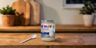 HiPP Gute Nacht Grießbrei pur im Glas auf einem Holztisch, mit Babylöffel davor in einer warm beleuchteten Küche