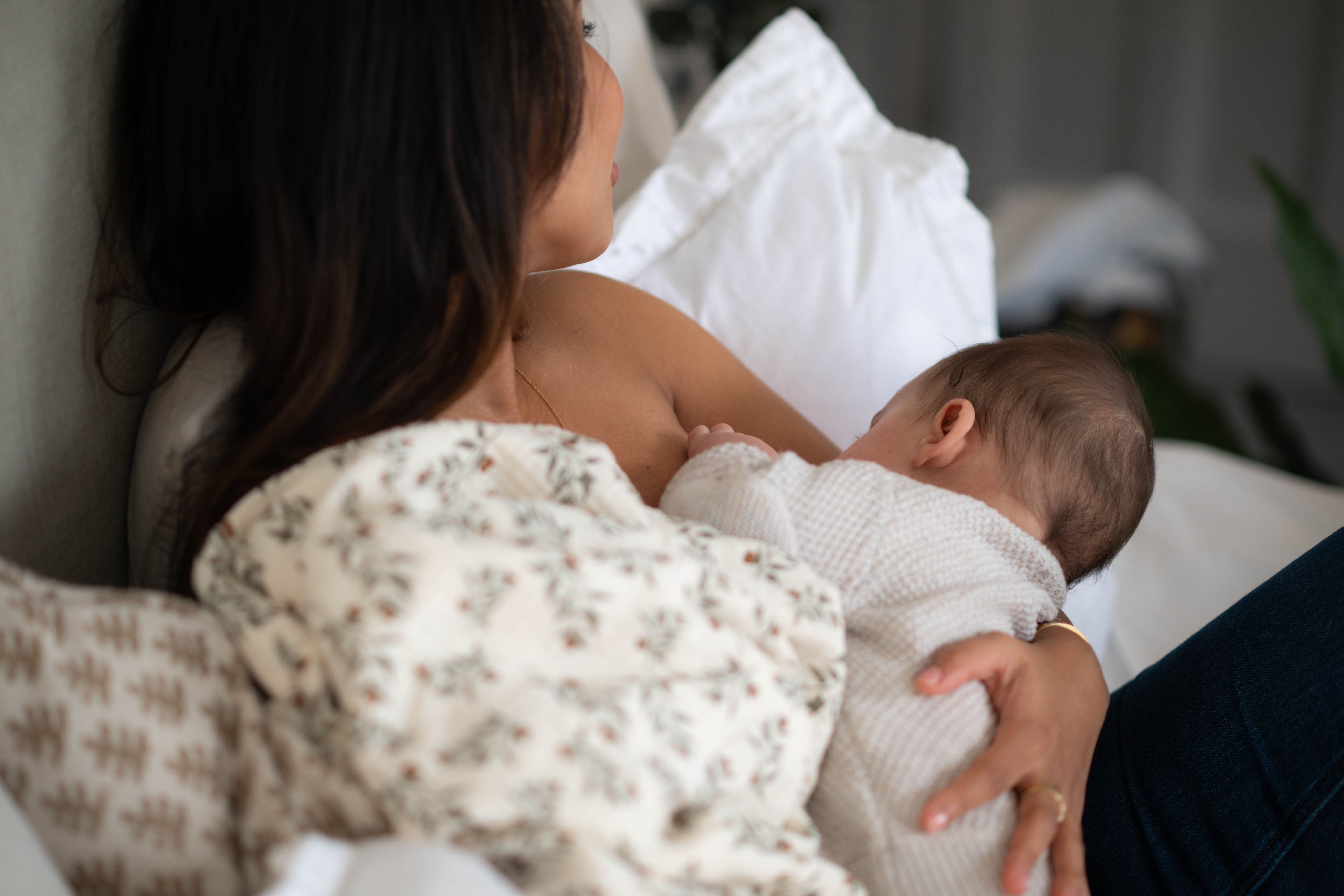 Mother breastfeeding her baby in bed, holding them close while resting against soft white pillows.