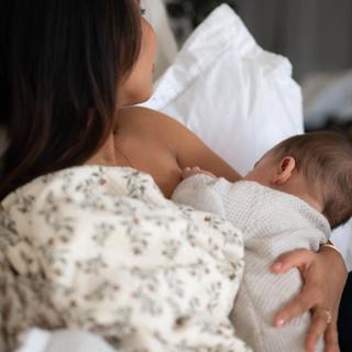 Mother breastfeeding her baby in bed, holding them close while resting against soft white pillows.