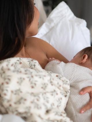 Mother breastfeeding her baby in bed, holding them close while resting against soft white pillows.