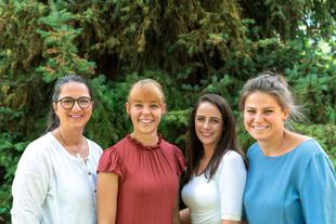 Vier Frauen stehen nebeneinander vor einem großen Nadelbaum und lächeln in die Kamera.