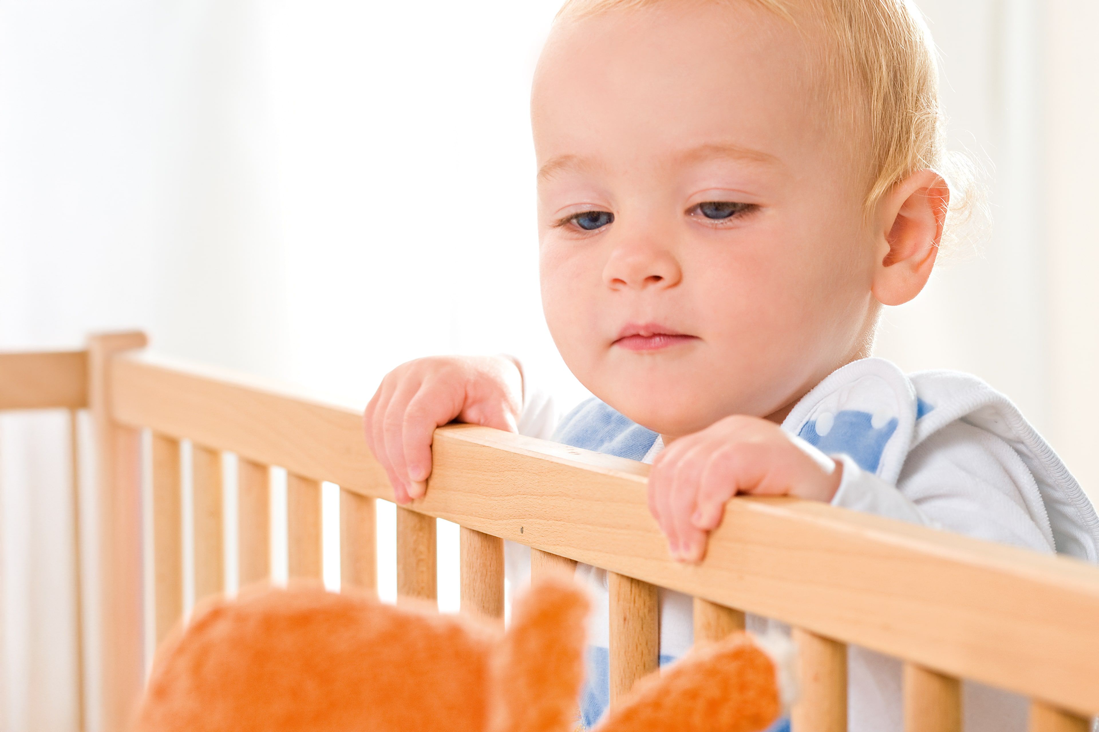 Ein kleines Kind, vermutlich ein Baby, hält sich mit konzentriertem Gesichtsausdruck an den Holzstäben eines Kinderbettes fest.