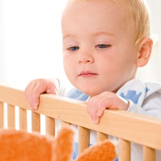 Ein kleines Kind, vermutlich ein Baby, hält sich mit konzentriertem Gesichtsausdruck an den Holzstäben eines Kinderbettes fest.