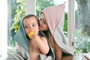 Ein kleines Kind, vermutlich ein Baby, eingewickelt in ein Handtuch, kaut auf einem gelben Gegenstand, während ein junges Mädchen, ebenfalls in ein Handtuch eingewickelt, sich vorbeugt, um dem Baby etwas ins Ohr zu flüstern.
