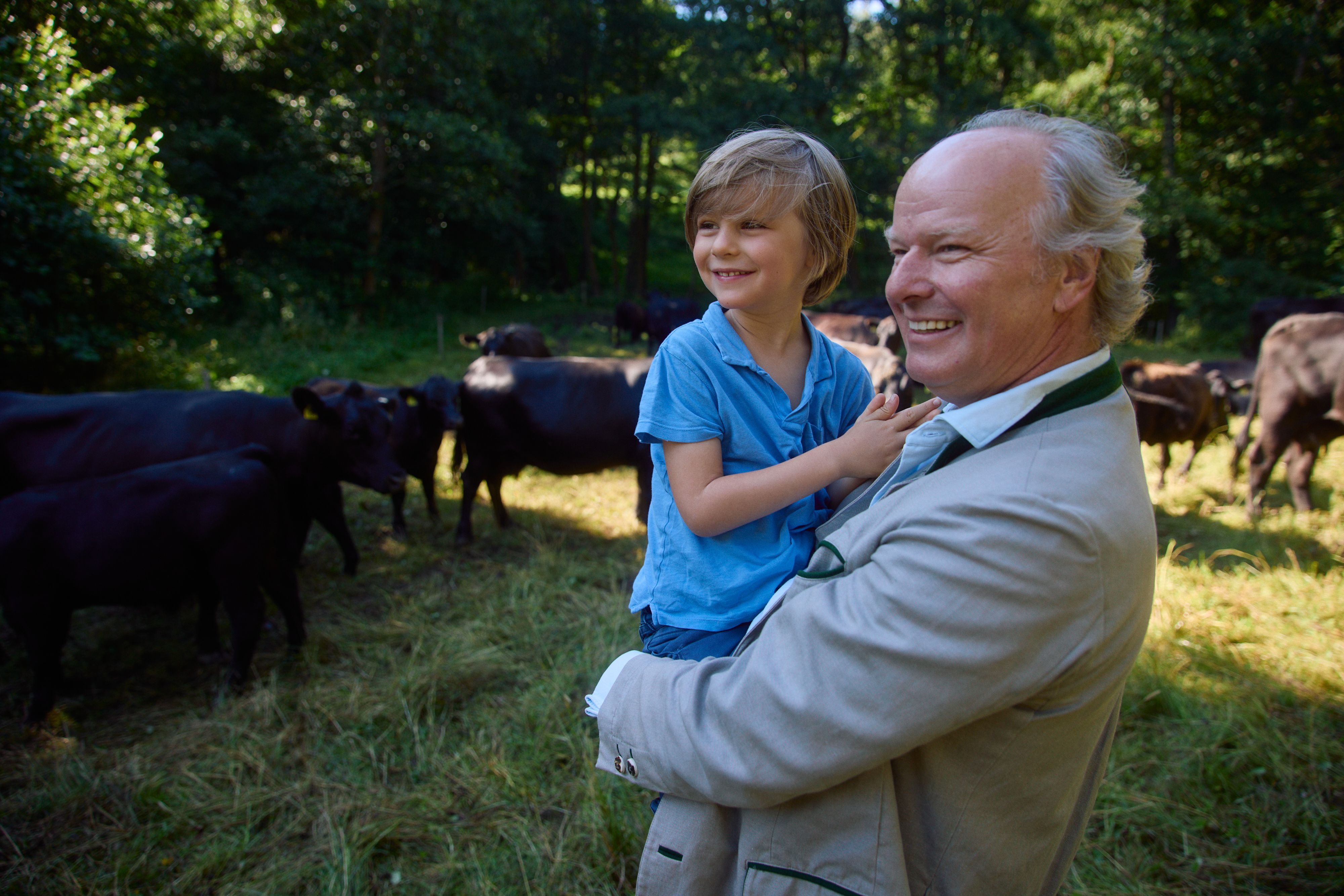 Großvater hält lächelnd sein Enkelkind auf einer Weide mit Rindern – generationsverbundene, naturnahe Familiengeschichte.