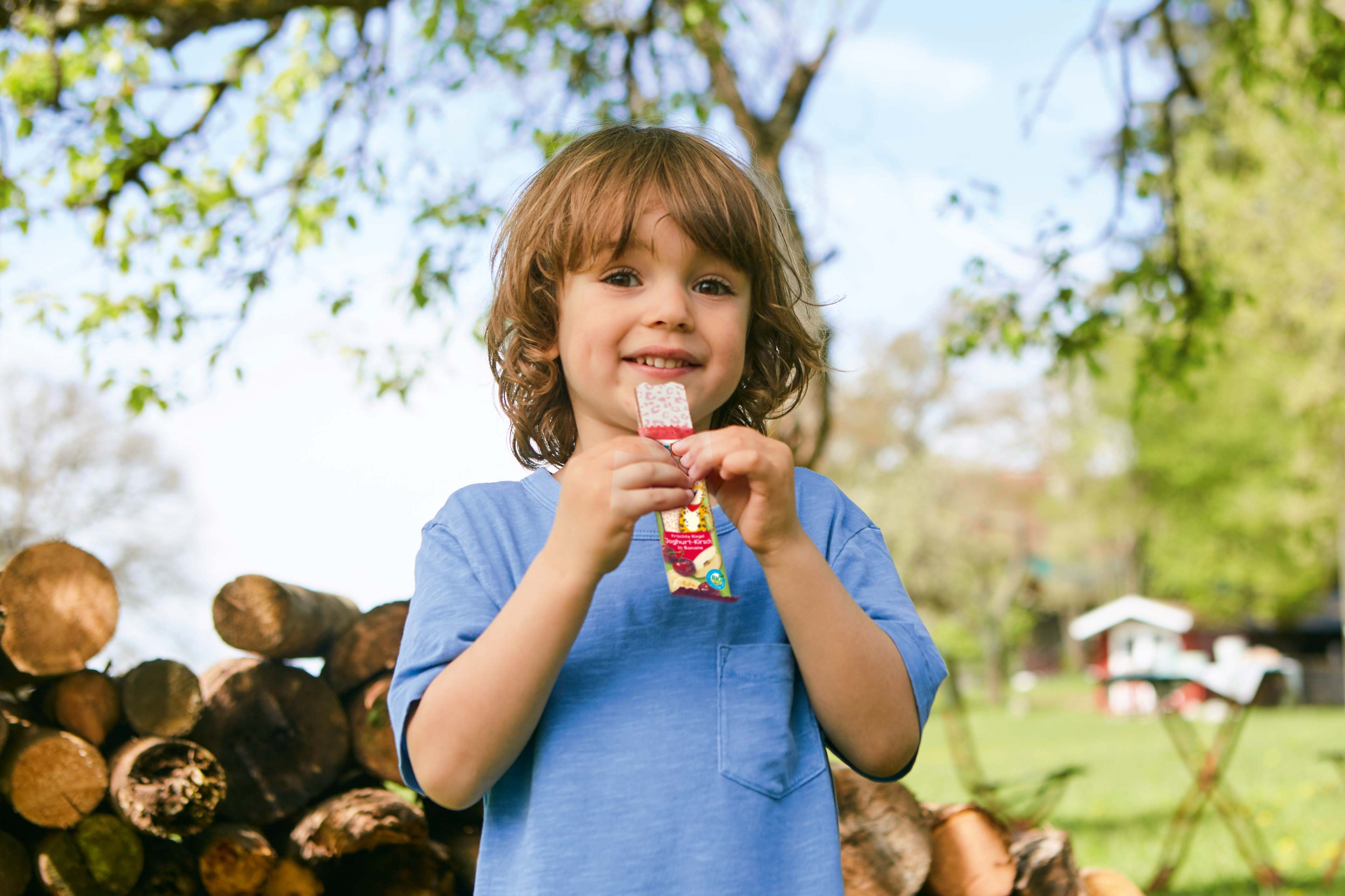 Lächelndes Kleinkind im Garten, das einen fruchtigen Kindersnack in der Hand hält.