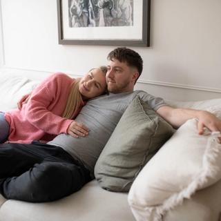 A couple relaxes on a cream-colored couch; the woman in a pink sweater lovingly rests her head on the man's chest while gently touching his stomach. They appear calm and content, enjoying a quiet moment together at home.