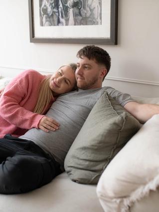 A couple relaxes on a cream-colored couch; the woman in a pink sweater lovingly rests her head on the man's chest while gently touching his stomach. They appear calm and content, enjoying a quiet moment together at home.
