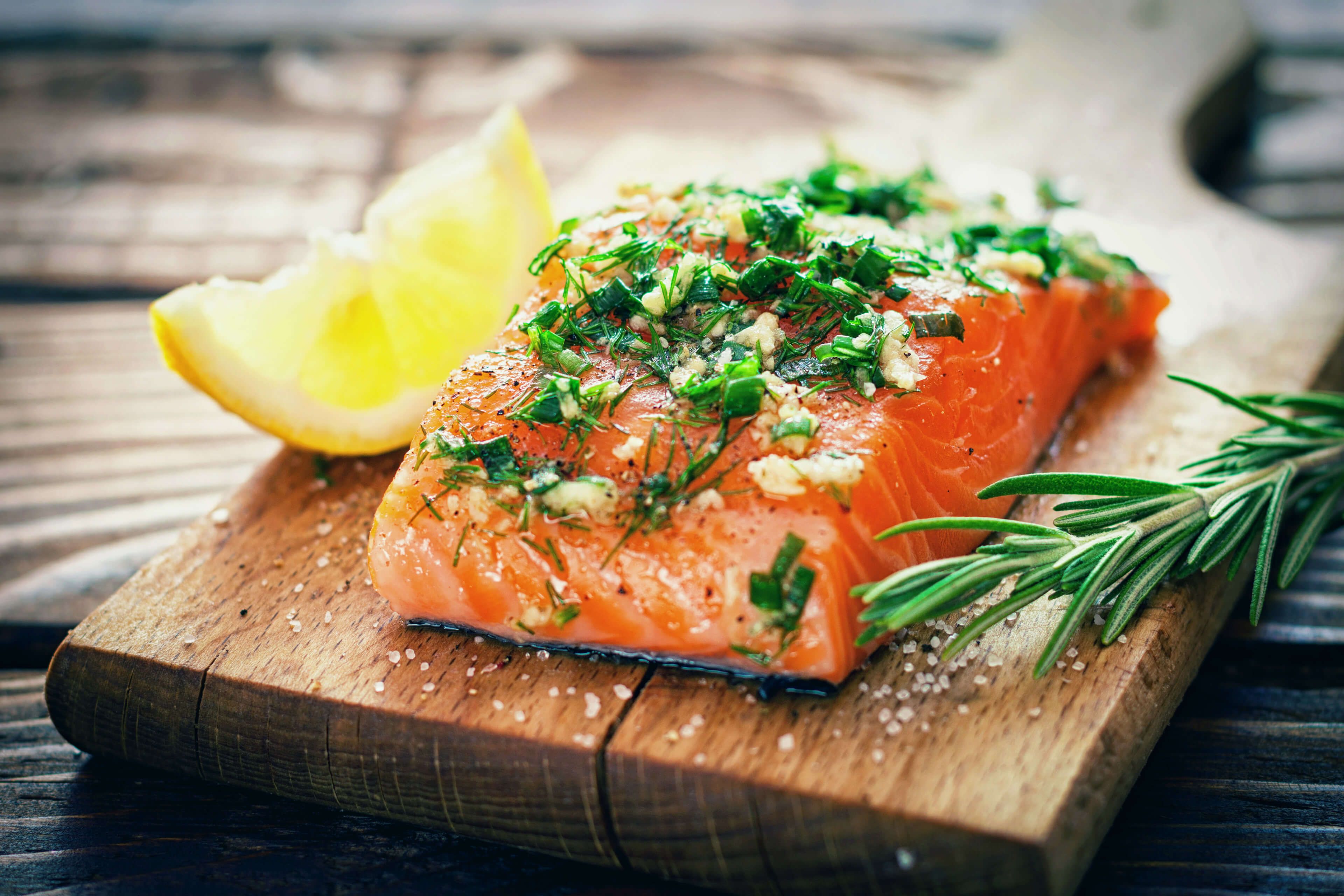 Ein Stück Lachs, garniert mit grünen Kräutern und einer Zitronenscheibe, auf einer Holzunterlage platziert.