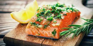 Ein Stück Lachs, garniert mit grünen Kräutern und einer Zitronenscheibe, auf einer Holzunterlage platziert.