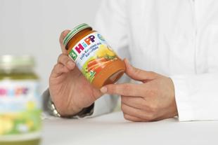 Person im weißen Kittel hält ein HiPP Bio-Babygläschen mit Frühkarotten und Kartoffeln vor hellem Hintergrund