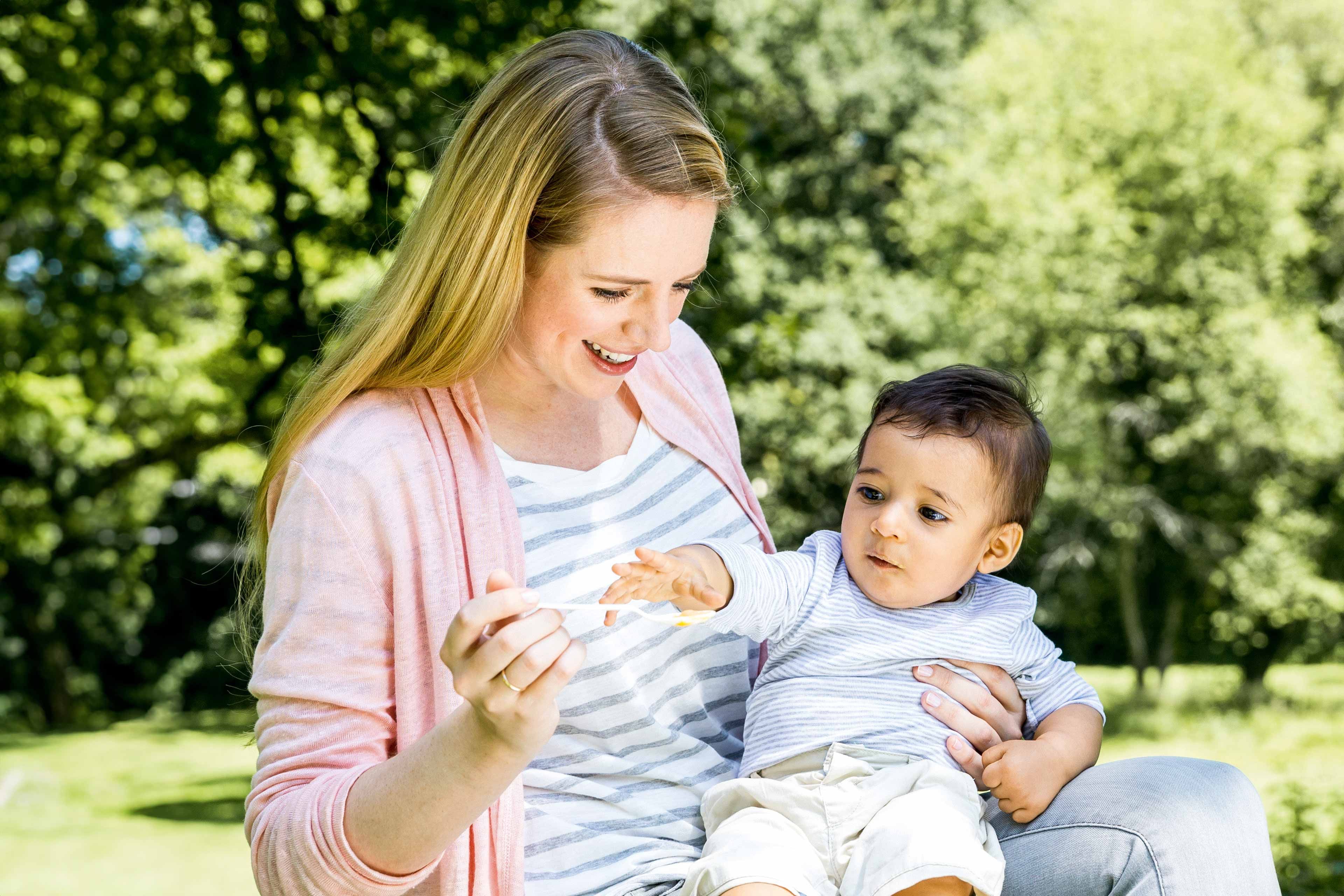 Eine Frau hält ein Baby im Freien, das sie mit einem Löffel füttert.