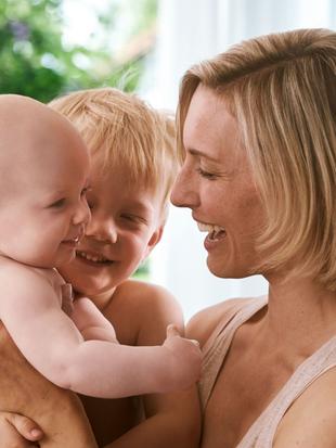 Ein freudiger Moment zwischen einer Mutter und ihren beiden Kindern, wobei die Mutter ein Baby im Arm hält und ein kleiner Junge sie anschaut.