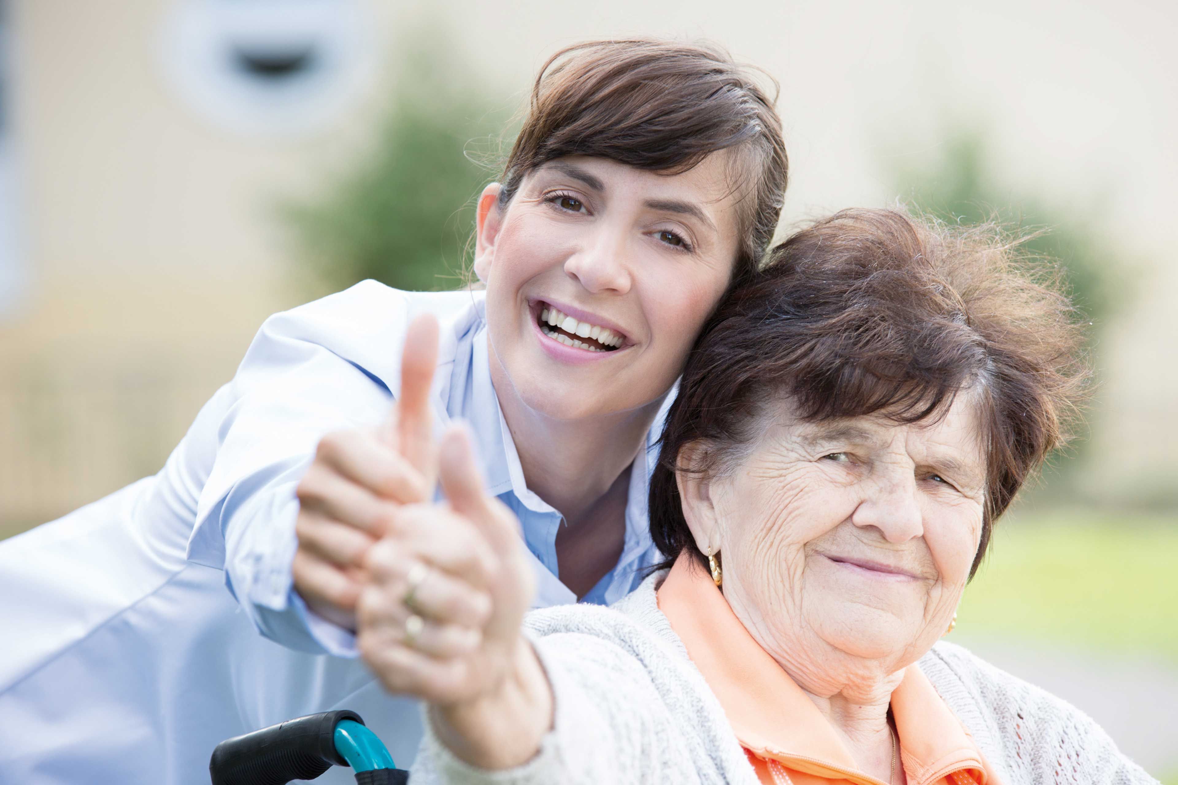 Jüngere Frau und Seniorin lächeln in die Kamera und zeigen gemeinsam den Daumen hoch als positives Erfahrungszeichen.
