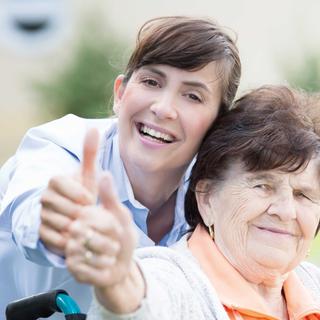 Jüngere Frau und Seniorin lächeln in die Kamera und zeigen gemeinsam den Daumen hoch als positives Erfahrungszeichen.