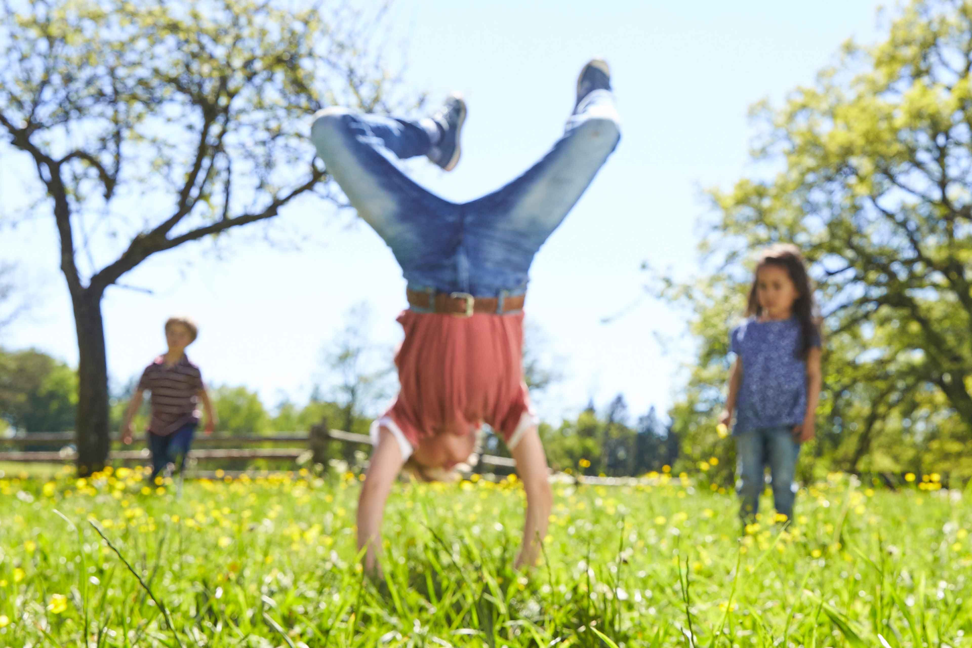 Ein fröhlicher Moment im Freien mit einem Kind, das einen Handstand inmitten eines Blumenfeldes macht, während zwei andere Kinder zusehen.