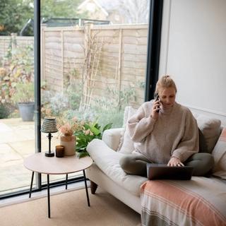 A woman sits cross-legged on a cozy beige sofa with a laptop in her lap, smiling as she talks on the phone. She’s wearing a chunky knit sweater and olive leggings, working from home in a bright living room with large glass doors that open to a lush garden. A small round table beside her holds a lamp, a candle, and a vase of soft pink flowers, creating a calm and inviting workspace.