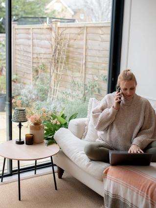 A woman sits cross-legged on a cozy beige sofa with a laptop in her lap, smiling as she talks on the phone. She’s wearing a chunky knit sweater and olive leggings, working from home in a bright living room with large glass doors that open to a lush garden. A small round table beside her holds a lamp, a candle, and a vase of soft pink flowers, creating a calm and inviting workspace.
