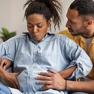 Pregnant woman experiencing labor contractions while sitting on a couch, supported by her partner who is holding her belly and offering comfort.