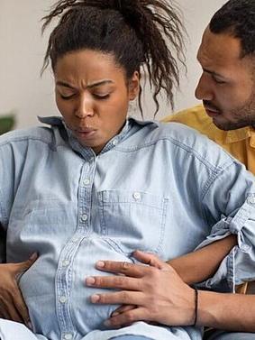 Pregnant woman experiencing labor contractions while sitting on a couch, supported by her partner who is holding her belly and offering comfort.