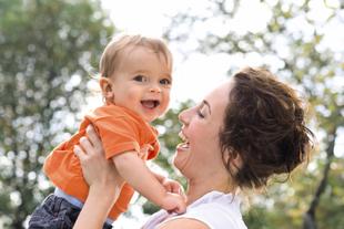 Lachende Mutter hebt ihr fröhliches Baby im Freien hoch, beide genießen gemeinsam die Zeit in der Natur.