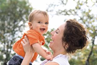 Lachende Mutter hebt ihr fröhliches Baby im Freien hoch, beide genießen gemeinsam die Zeit in der Natur.