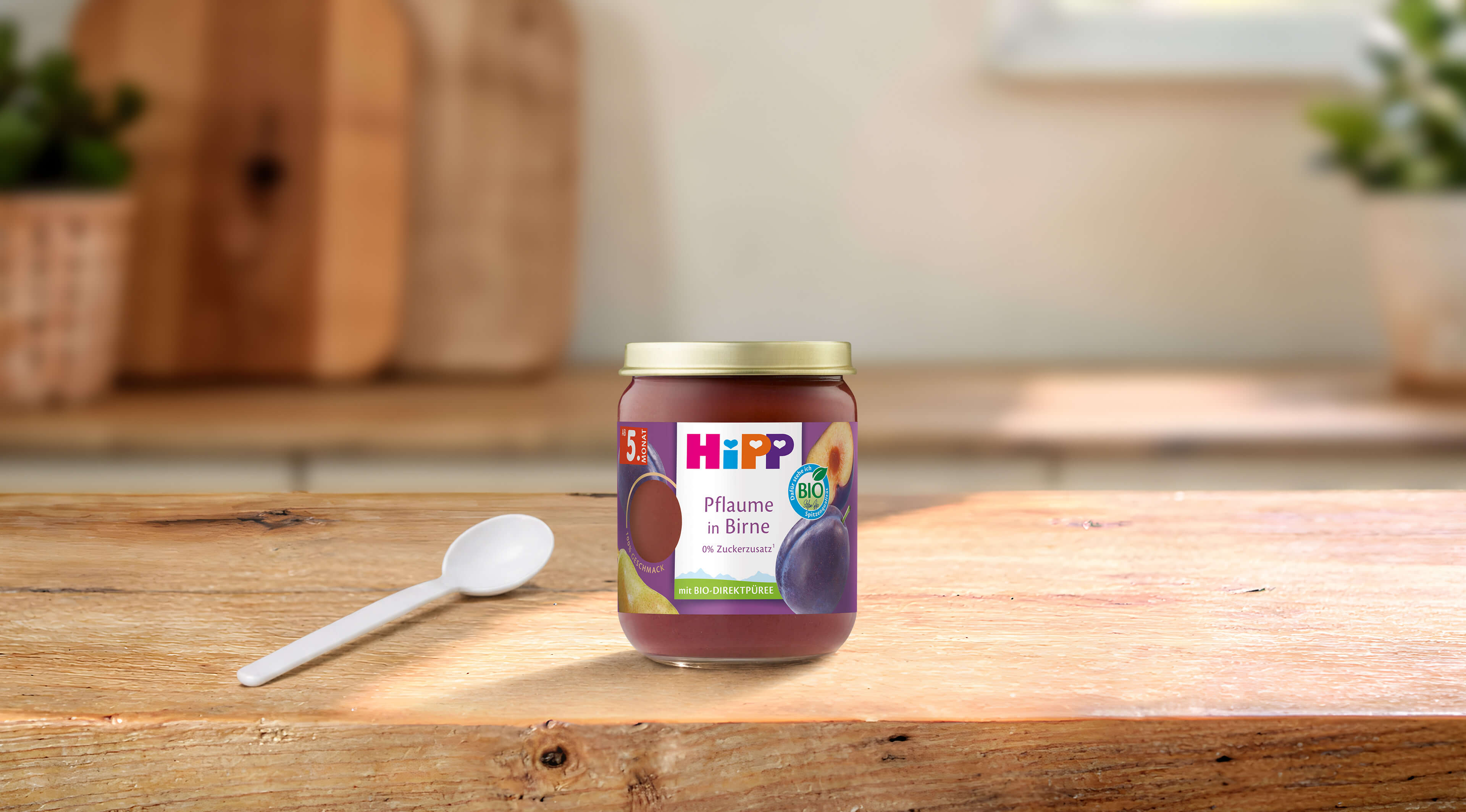 HiPP Bio Babyfruchtglas „Pflaume in Birne“ auf Holztisch mit Löffel, im hellen, gemütlichen Küchenhintergrund