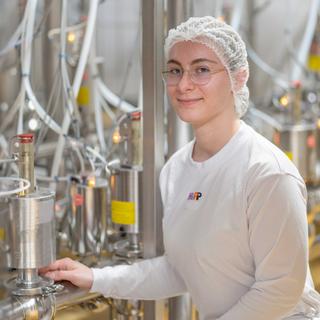 Junge Frau mit Haarnetz und Brille bedient in einer Produktionsanlage Edelstahlbehälter mit Schläuchen im Hintergrund