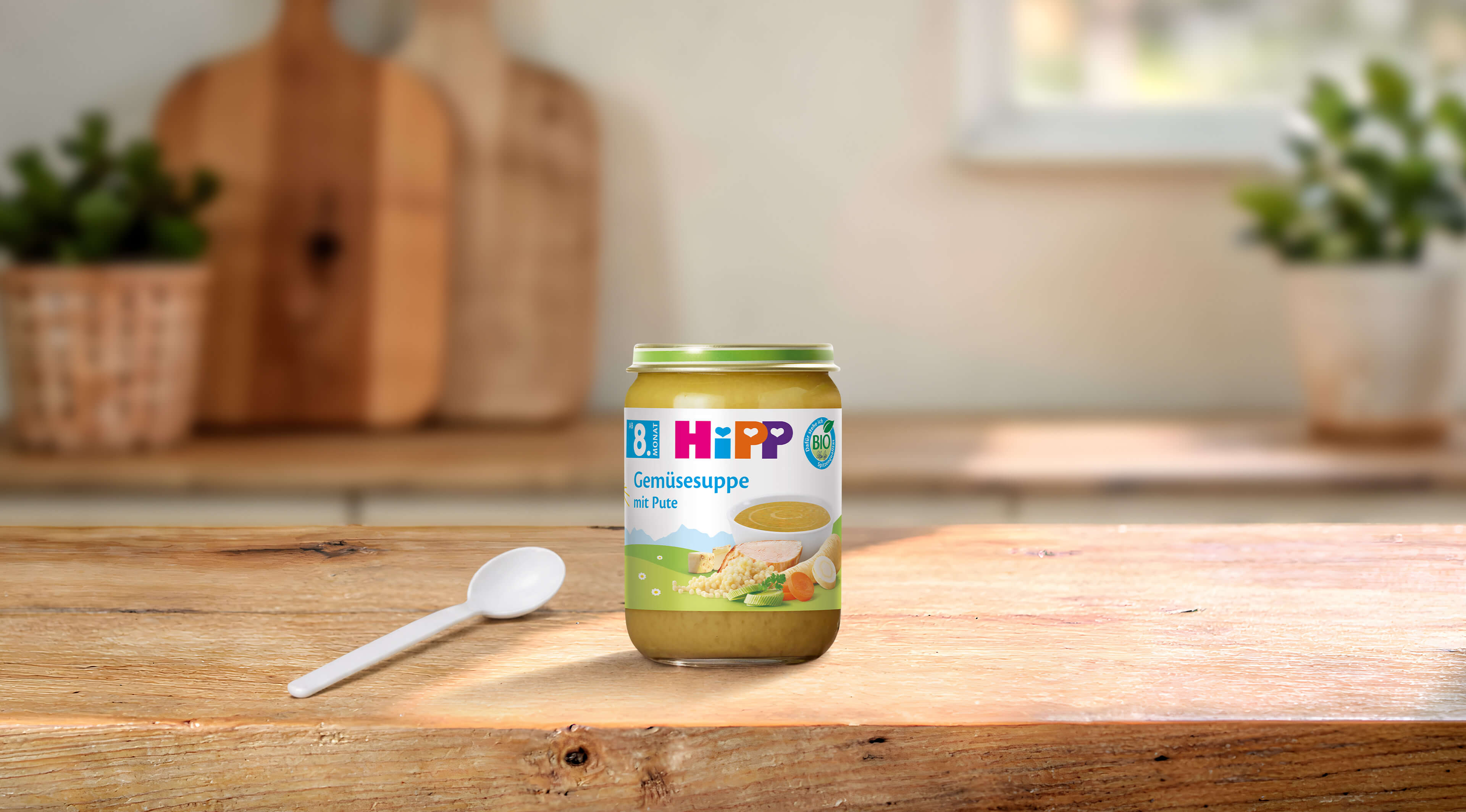 HiPP Bio-Glas Gemüsesuppe mit Pute ab dem 8. Monat, Glas mit Löffel auf Holztisch in heller Küche