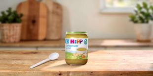 HiPP Bio-Glas Gemüsesuppe mit Pute ab dem 8. Monat, Glas mit Löffel auf Holztisch in heller Küche