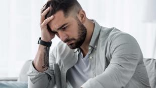 Ein Mann mit Bart, der ein helles Hemd trägt, hält seinen Kopf in der Hand und scheint in Gedanken versunken zu sein.