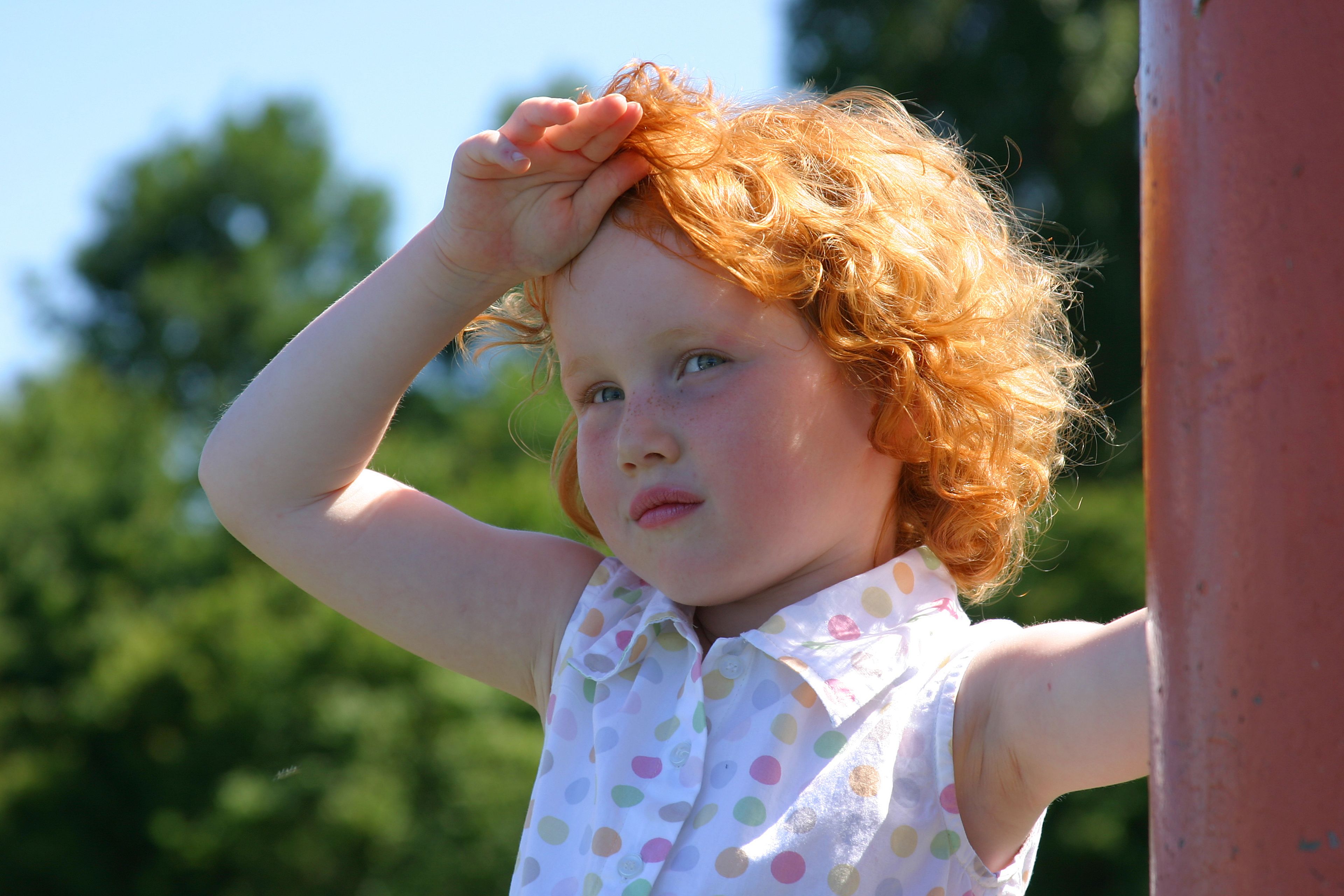 Ein junges Mädchen mit lockigem rotem Haar, das seine Augen mit der Hand vor der Sonne schützt, vor einem grünen Hintergrund.